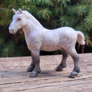 Puede incluir: Figura detallada de un caballo en posición de pie. El caballo es principalmente blanco con manchas y patas grises. El fondo es un follaje verde borroso, y el caballo está sobre una superficie de madera.