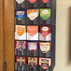 Puede incluir: Un organizador de bolsitas de té negro de varios niveles, lleno de varias marcas y sabores de té. Las bolsitas de té están en compartimentos individuales, con marcas como Twinings y Choice Organics. Montado en un armario de madera.