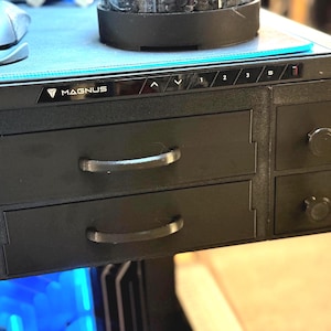 May include: Black desk with two drawers and two smaller compartments. The desk has a control panel with buttons and the word "MAGNUS" printed on it. A computer mouse is visible on the desk surface.