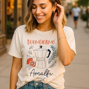 Può includere: T-shirt color crema con le parole italiane "Buongiorno Amalfi Italia" in rosso e blu navy. Il design include una caffettiera, fette d'arancia ed elementi botanici. La maglietta è realizzata in materiale morbido.