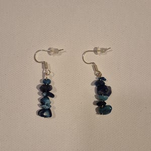 May include: A pair of dangle earrings featuring a stack of blue and teal-colored stones. The stones are of varying shapes and sizes, strung together with silver-colored wire and hooks. The earrings are displayed against a white background.