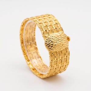 Pode incluir: Uma pulseira de tom dourado com uma banda larga e flexível, apresentando um padrão de elos retangulares repetitivos e um acabamento texturizado. A pulseira inclui um fecho central oval proeminente com um design de superfície cruzada.