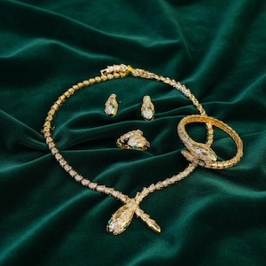 Könnte beinhalten: Ein goldfarbenes Schmuckset mit Schlangenmuster. Das Set umfasst eine Halskette, Ohrringe, einen Ring und ein Armband, die alle mit klaren Steinen und grünen Akzenten verziert sind. Der Schmuck wird auf einem dunkelgrünen Samtstoff präsentiert.