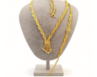 May include: A gold-colored necklace and bracelet set displayed on a gray mannequin. The necklace features a braided design with a tassel pendant. The bracelet has a similar braided pattern. The jewelry is presented against a white background.