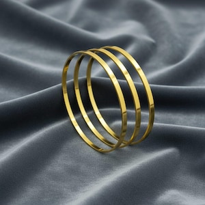 Könnte beinhalten: Vier goldfarbene Armreifen liegen auf einem grauen Samtstoff. Die Armreifen sind dünn und haben eine glatte, reflektierende Oberfläche. Der Schmuck wird in einem Studio-Setting präsentiert, mit weicher Beleuchtung, die den Glanz des Goldes hervorhebt.