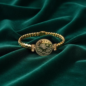 Könnte beinhalten: Ein goldfarbenes Armband mit einem münzartigen Mittelstück. Das Armband hat ein kettenartiges Design und einen Verschluss. Die Münze zeigt ein Emblem. Das Armband wird auf einer dunkelgrünen Samtoberfläche präsentiert.