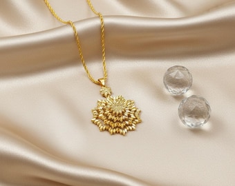 May include: A gold-colored necklace with a floral pendant and a delicate chain. The pendant has a layered design and is displayed on a cream-colored satin surface. Two clear, faceted crystal spheres are next to the necklace.