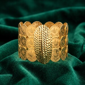 May include: A wide gold cuff bracelet composed of multiple rows of circular gold coins featuring intricate engravings and a central, large, oval-shaped gold plaque with a highly textured, diamond-cut geometric pattern across its surface.