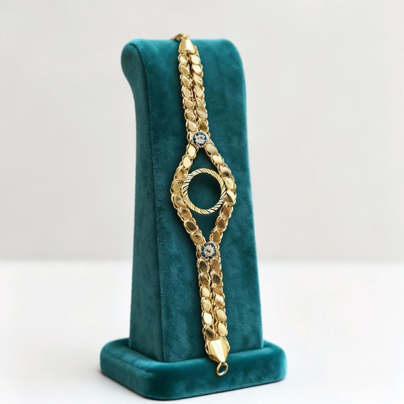 Puede incluir: Una pulsera dorada con un dise&ntilde;o &uacute;nico, con dos cadenas paralelas conectadas por un elemento circular central y adornada con peque&ntilde;os detalles azules. La pulsera se exhibe sobre un soporte de terciopelo turquesa, mostrando sus intrincados detalles.