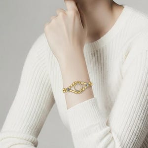 Puede incluir: Pulsera dorada con un dise&ntilde;o ovalado de centro abierto. La pulsera presenta un patr&oacute;n texturizado de cuentas y peque&ntilde;os detalles azules. La joya se lleva en una mu&ntilde;eca, contra un su&eacute;ter blanco.