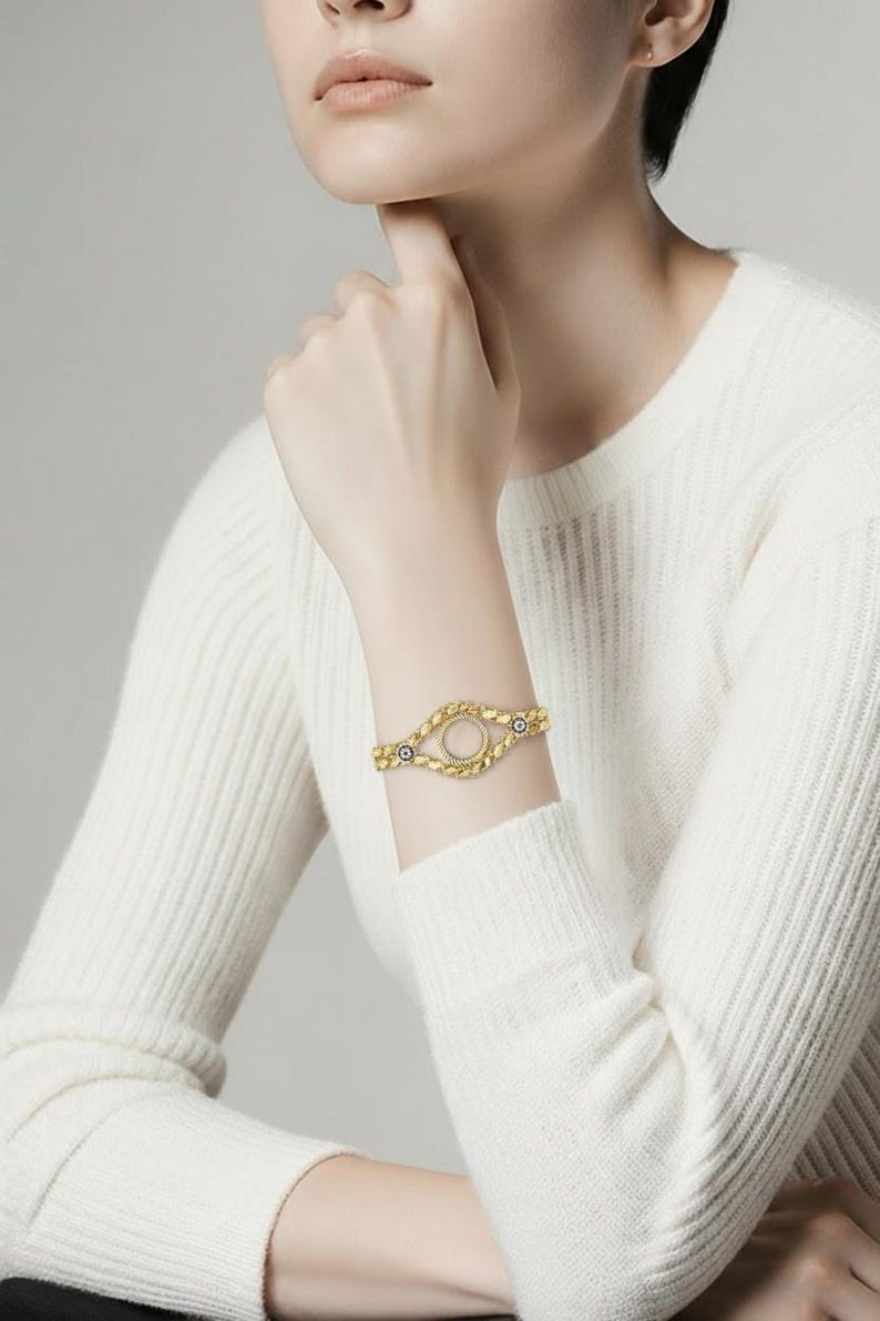 Puede incluir: Pulsera dorada con un dise&ntilde;o ovalado de centro abierto. La pulsera presenta un patr&oacute;n texturizado de cuentas y peque&ntilde;os detalles azules. La joya se lleva en una mu&ntilde;eca, contra un su&eacute;ter blanco.