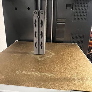 Könnte beinhalten: Ein schwarzes, 3D-gedrucktes Objekt, das einem Butterflymesser ähnelt, steht auf einem goldfarbenen Druckbett. Das Objekt hat ein symmetrisches Design mit rautenförmigen Aussparungen. Der Drucker ist schwarz mit silbernen Stäben.