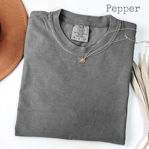 Op de afbeelding: Een gevouwen, peperkleurig t-shirt met een ronde hals en een klein label. Een zilveren ketting met een hanger ligt op het shirt. Een bruine hoed en gedroogde plantenstengels zijn op de achtergrond. Het woord "Pepper" staat rechtsboven.