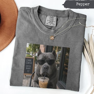 May include: A pepper-coloured crew neck t-shirt featuring a graphic print of a Cane Corso dog wearing black sunglasses and holding a plastic cup with a straw. The shirt has a Comfort Colors label at the inner neckline and a thin gold-toned necklace.