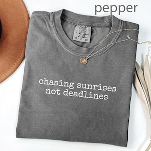May include: A grey t-shirt with the text "chasing sunrises not deadlines" in white. A gold necklace with a pendant is draped on the shirt. A brown hat and dried plant are in the background.