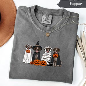 Op de afbeelding: Een peperkleurig t-shirt met een afbeelding van vier honden in Halloween-kostuums. Eén hond is een geest, één een heks, één een mummie en één een vampier. Ze worden omringd door oranje pompoenen en een kleine grafsteen met het opschrift RIP.