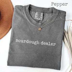 May include: A gray t-shirt with the text "sourdough dealer" in white. A gold necklace with a pendant rests on the shirt. The shirt is folded and next to a brown hat and dried plant.