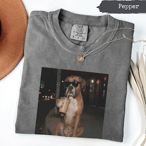 Può includere: Una t-shirt a girocollo color pepe con una stampa grafica di un cane boxer marrone che indossa occhiali da sole neri e tiene in mano un bicchiere di plastica con una cannuccia. Presenta un'etichetta Comfort Colors sul collo e una catenina d'oro.