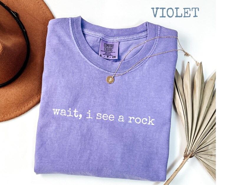 May include: A violet Comfort Colors t-shirt with the text "wait, i see a rock" in white. A gold necklace with a pendant is draped on the shirt. A brown hat is visible in the upper left corner.