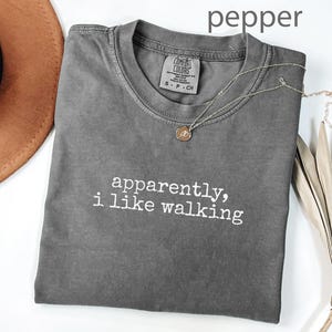 Puede incluir: Una camiseta gris con el texto "apparently, i like walking" en blanco. Un collar dorado con un pequeño colgante descansa sobre la camiseta. La camiseta está doblada y junto a un sombrero marrón y una planta seca.