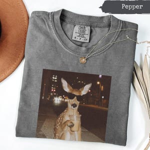 May include: A pepper-coloured crew neck t-shirt featuring a graphic print of a deer wearing black sunglasses and holding a clear plastic cup with a straw. The shirt has a Comfort Colors brand label on the inside collar with size S P CH printed on it.