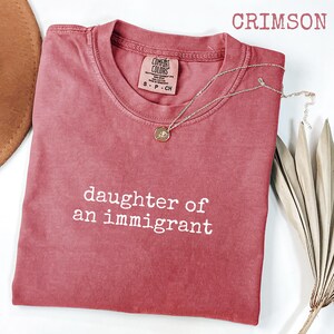 May include: Crimson-colored t-shirt with the text "daughter of an immigrant" in white. A gold necklace with a pendant rests on the shirt. The shirt is a Comfort Colors brand. A brown hat and dried plant are in the background.