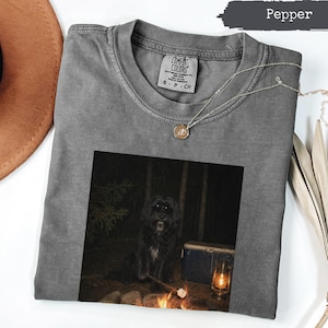 Pode incluir: Uma t-shirt cor de pimenta com uma impressão gráfica de um cão preto sentado junto a uma fogueira à noite, com uma geleira azul e uma lanterna por perto. A t-shirt tem uma etiqueta da marca Comfort Colors e gola redonda canelada.