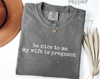 Puede incluir: Camiseta color pimienta con el texto "be nice to me my wife is pregnant" en blanco. Un collar dorado con un colgante está colocado sobre la camiseta. La camiseta está doblada sobre una superficie blanca.