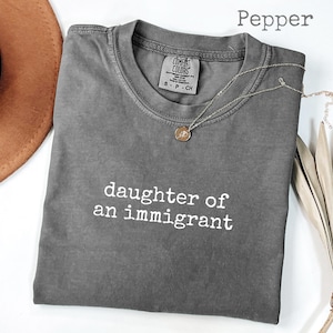 May include: A gray Comfort Colors t-shirt with the text "daughter of an immigrant" in white. A gold pendant necklace and a brown hat are also visible. The shirt is a size small.