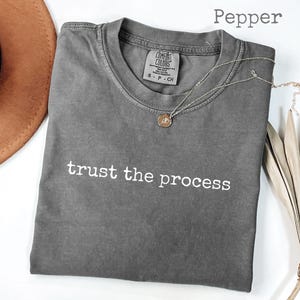 May include: A gray Comfort Colors t-shirt with the phrase "trust the process" in white. A gold pendant necklace and a brown hat are next to the shirt. The shirt is folded and has a tag on the collar.