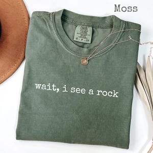 May include: A moss green t-shirt with the text "wait, i see a rock" in white. A gold necklace with a pendant is draped across the shirt. A brown hat and dried plant are in the background. The shirt has a crew neck and short sleeves.