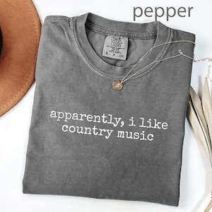 May include: A gray t-shirt with the text "apparently, i like country music" in white. A gold necklace with a pendant is on the shirt. A brown hat and dried plant are in the background.