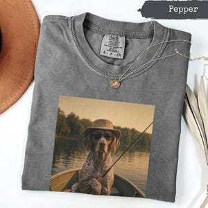 Puede incluir: Una camiseta gris carbón de cuello redondo con un estampado gráfico de un perro con sombrero de pescador y gafas de sol, sentado en un barco con una caña de pescar. La prenda incluye una etiqueta de Comfort Colors en el cuello.
