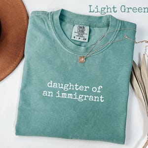 May include: Light green t-shirt with the text "daughter of an immigrant" in white. A gold necklace with a pendant rests on the shirt. The shirt is folded and next to a brown hat and dried plant.