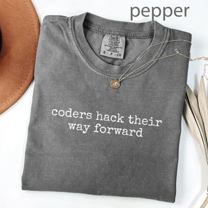Può includere: Una maglietta grigia con la scritta bianca "coders hack their way forward". Una collana d'oro con un ciondolo è sulla maglietta. La maglietta è piegata, accanto a un cappello marrone e una pianta secca.