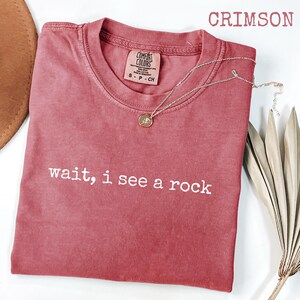 May include: Crimson-colored t-shirt with the text "wait, i see a rock" in white. A silver necklace with a pendant rests on the shirt. The shirt is a Comfort Colors brand, and a brown hat is partially visible.