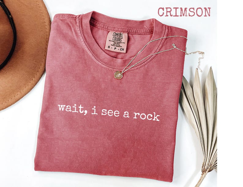 May include: Crimson-colored t-shirt with the text "wait, i see a rock" in white. A silver necklace with a pendant rests on the shirt. The shirt is a Comfort Colors brand, and a brown hat is partially visible.