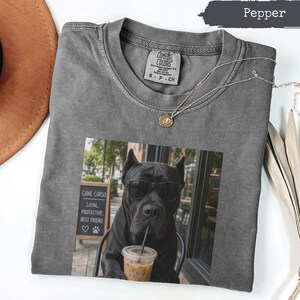 Könnte beinhalten: Ein pfefferfarbenes T-Shirt mit einem Aufdruck eines schwarzen Cane Corso Hundes, der eine Sonnenbrille trägt und einen Eiskaffee trinkt. Der Hund steht neben einem Schild mit der Aufschrift Cane Corso, Loyal, Protective, Best Friend. Eine goldene Kette liegt auf.