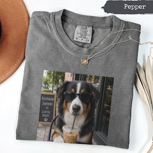 Könnte beinhalten: Ein pfeffergraues T-Shirt mit Rundhalsausschnitt und einem Aufdruck eines Australian Shepherd Hundes, der eine schwarze Sonnenbrille trägt und aus einem durchsichtigen Plastikbecher mit Strohhalm trinkt. Ein Schild zeigt Australian Shepherd Coffee Buddy.