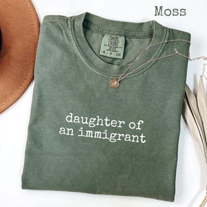 May include: A moss green t-shirt with the text "daughter of an immigrant" in white. A gold necklace with a small pendant rests on the shirt. A brown hat and dried plant are in the background.
