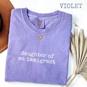 May include: A violet Comfort Colors t-shirt with the text "daughter of an immigrant" in white. A gold necklace with a pendant is draped over the shirt. A brown hat and dried plant are in the background.