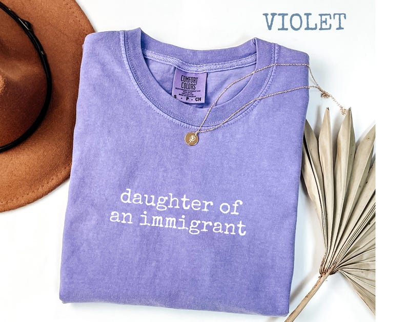 May include: A violet Comfort Colors t-shirt with the text "daughter of an immigrant" in white. A gold necklace with a pendant is draped over the shirt. A brown hat and dried plant are in the background.