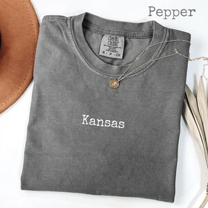 May include: A gray t-shirt with the word "Kansas" embroidered in white. A gold pendant necklace rests on the shirt. The word "Pepper" is written in the top right corner.