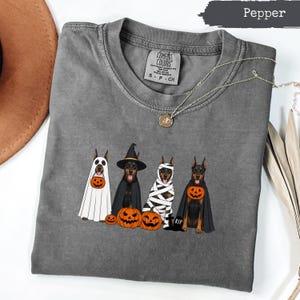 May include: A charcoal gray t-shirt featuring a graphic of four Doberman dogs dressed in Halloween costumes, including a ghost, a witch, a mummy, and a vampire, accompanied by orange jack-o'-lantern pumpkins and a small tombstone labeled RIP.