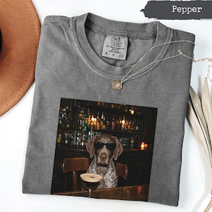 Può includere: Una t-shirt color pepe con una stampa grafica di un cane con occhiali da sole, seduto al bancone di un bar con un bicchiere da martini. La maglietta ha un girocollo e un'etichetta Comfort Colors all'interno. Una delicata collana dorata è appoggiata sopra.