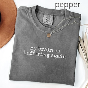 May include: A gray t-shirt with the text "my brain is buffering again" in white. A gold necklace with a pendant is on the shirt. The shirt is folded on a white surface. A brown hat is visible in the upper left corner.