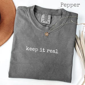 May include: A gray Comfort Colors t-shirt with the phrase "keep it real" in white. A gold pendant necklace rests on the shirt. A brown hat and dried palm leaves are in the background. The shirt is labeled "Pepper".