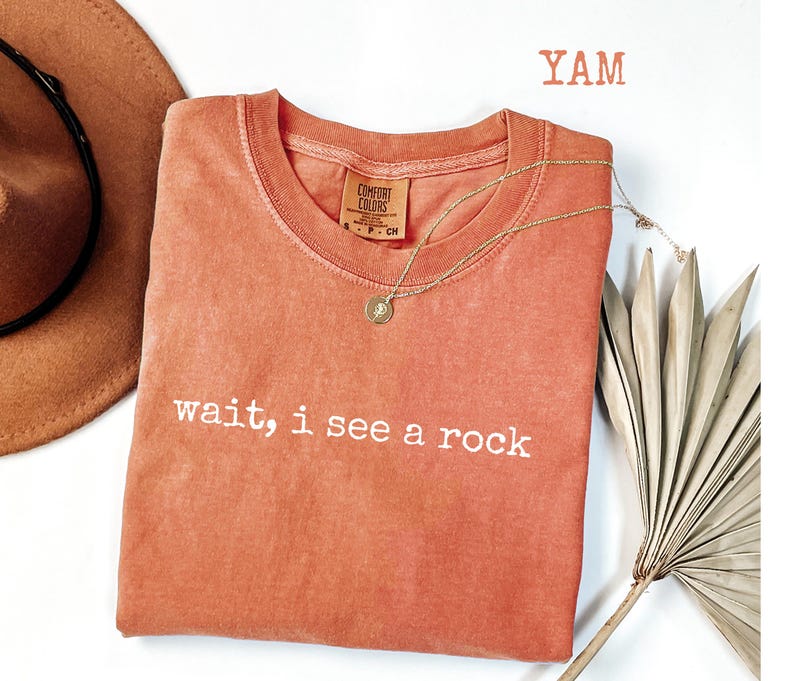 May include: A coral-colored t-shirt with the text "wait, i see a rock" in white. A gold necklace with a pendant is on the shirt. A brown hat and a dried palm leaf are in the background. The word "YAM" is at the top.