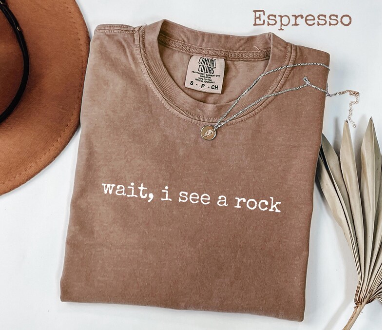 May include: Espresso-colored t-shirt with the text "wait, i see a rock" in white. A silver necklace with a small pendant is draped across the shirt. The shirt is a Comfort Colors brand.