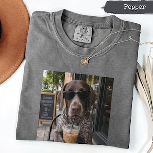 Può includere: Una t-shirt girocollo grigio antracite con una stampa grafica di un bracco tedesco a pelo corto che indossa occhiali da sole neri e beve da un bicchiere di plastica con una cannuccia. La maglia ha un'etichetta Comfort Colors sul collo.